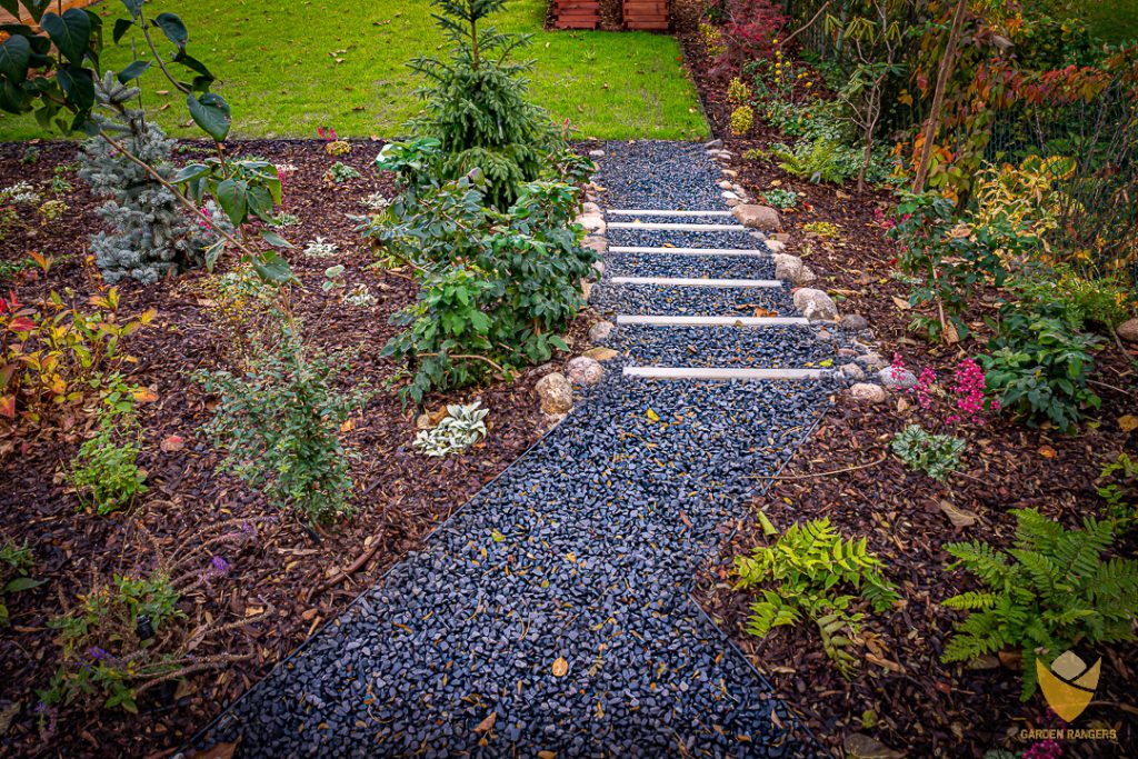 Geometryczna ścieżka w ogrodzie wykonana z drobnego grysu bazaltowego, ze stopniami stworzonymi z płyt betonowych. Ścieżka przechodzi wśród rabat, prowadząc na trawnik.